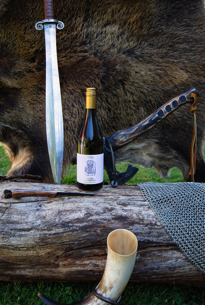 Halvard Alter Schwede Weißwein von Weingut Schwedhelm Flasche auf Baumstamm mit Schwert, Axt und Wikinger Horn