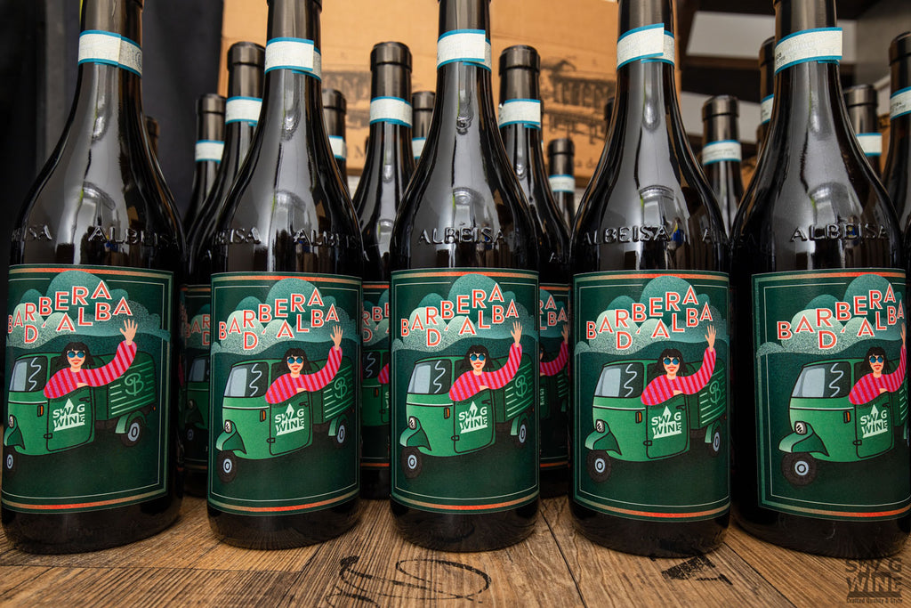 Mehrere Flaschen Barbera d'Alba 2015 von Swagwine mit Frau winkend aus einem Piaggio Ape