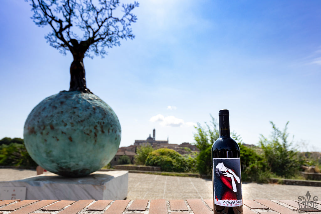 Concalma - Rotwein aus der Toskana Flasche vor Statue eines Baums der aus der Erdkugel wächst vor Stadtkulisse