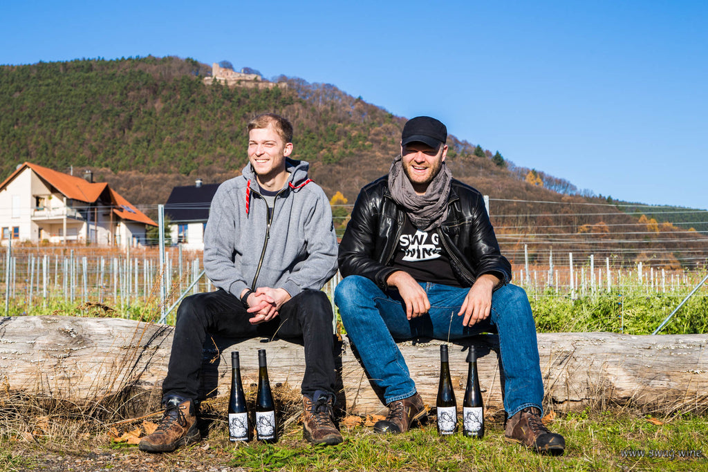 Winzer Georg Meier und Sven von Swagwine sitzen auf einem Baumstamm mit Weinflaschen auf Boden